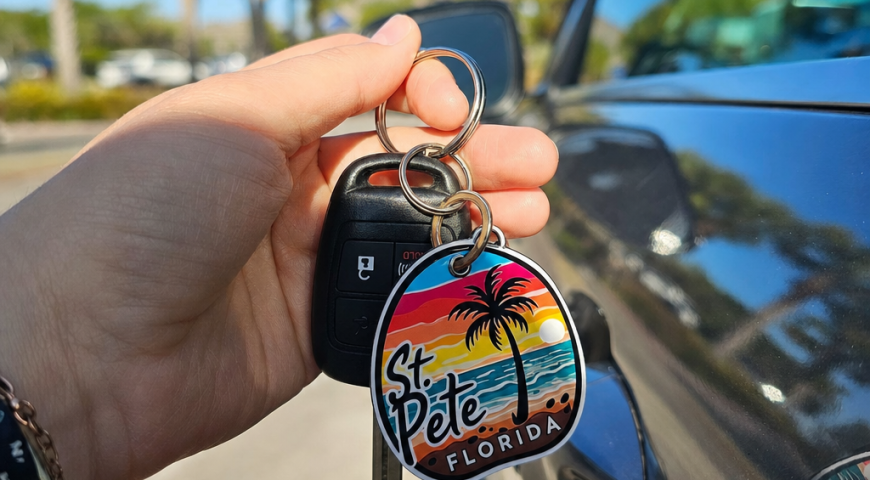 Keychain reading 'St Pete Florida' with a palm tree and sunset, held with car keys beside a car door edge, sunny day layout info accessory view
